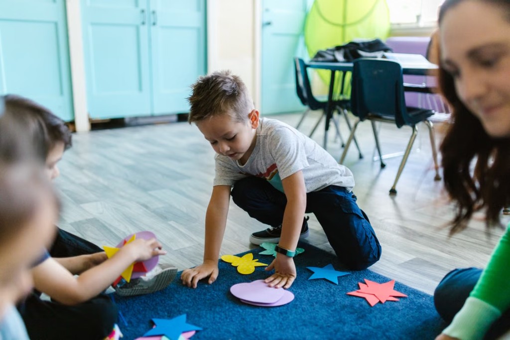 A young boy sorting colorful paper shapes on a blue rug with a teacher nearby